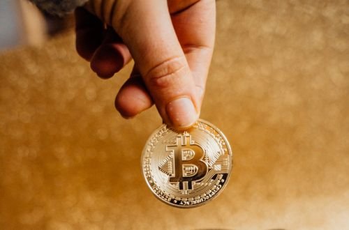 A hand places a Bitcoin into a piggy bank labeled 'Crypto', symbolizing savings and investment.