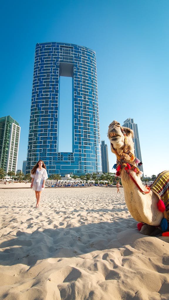 A woman walks on a Dubai beach with a camel and modern skyscrapers in the background.
