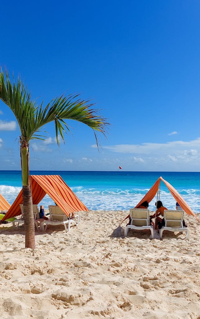 Beach scene in Cancún with orange tents, palm tree, and clear blue water. Perfect summer getaway.