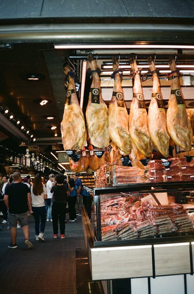 Hanging Spanish jamon in a bustling Barcelona market with customers browsing the meat section.