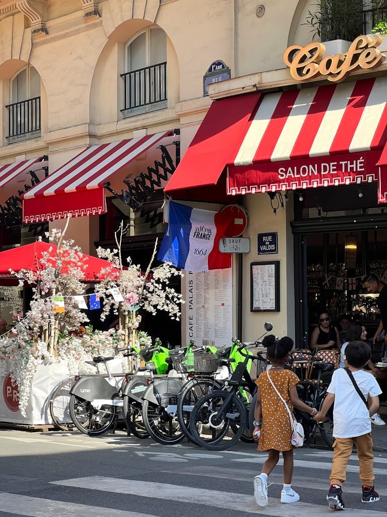Parisian café with bicycles, children, and red-striped awnings on a city street in daylight.