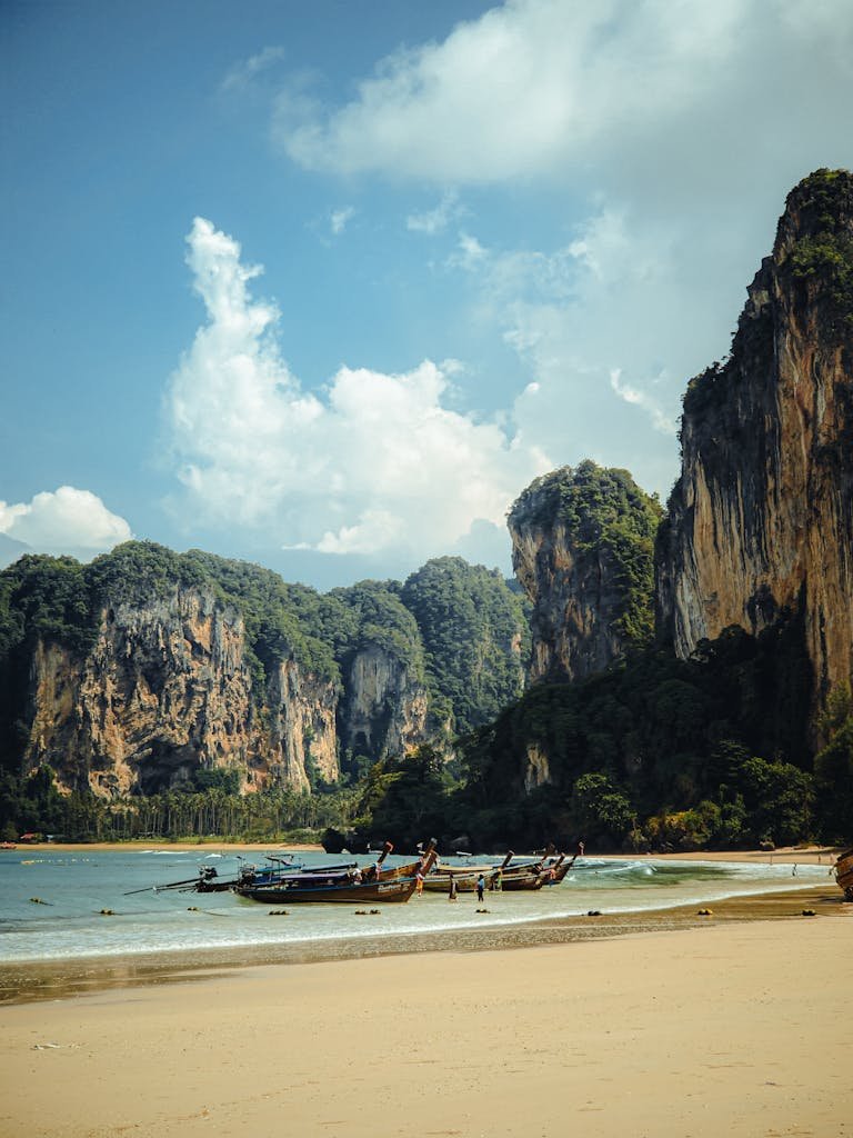 Serene beach with towering cliffs in Krabi, Thailand, showcasing traditional longtail boats under a clear sky.
