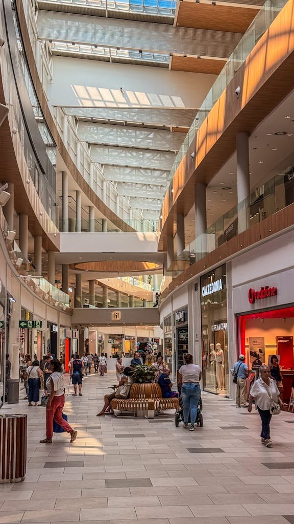 Spacious shopping mall interior with people walking, stores, and natural sunlight.