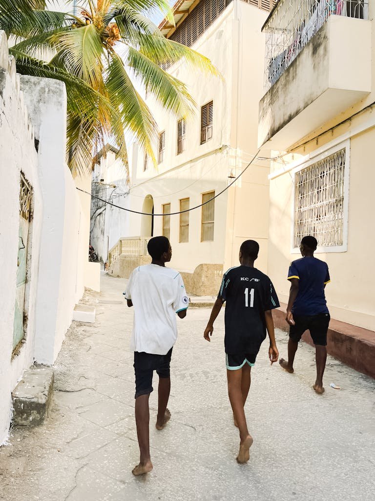 Three teenagers walk down a sunlit urban street in Tanzania with palm trees and buildings surrounding them.