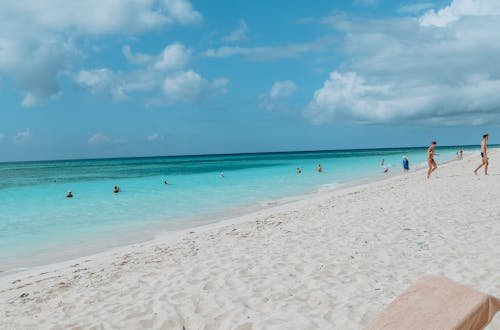 From above of sun bed with towel and ripe pineapple on sandy sea shore with unrecognizable tourists under blue cloudy sky