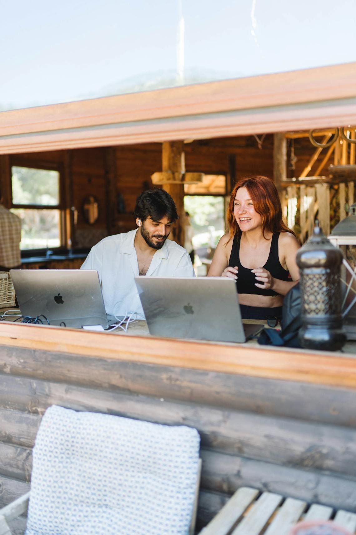 Two young adults working on laptops in a cozy, wooden interior with natural light.